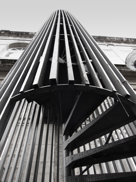 black and white photograph, portrait layout, looking up a set of outdoor spiral steps with vertical strips surrounding the steps