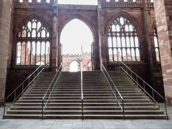 colour photograph of stone steps leading from bottom to top, with arched  bombed out stained glass windwos on either side and an oped doorway leading to the bombed cathedral