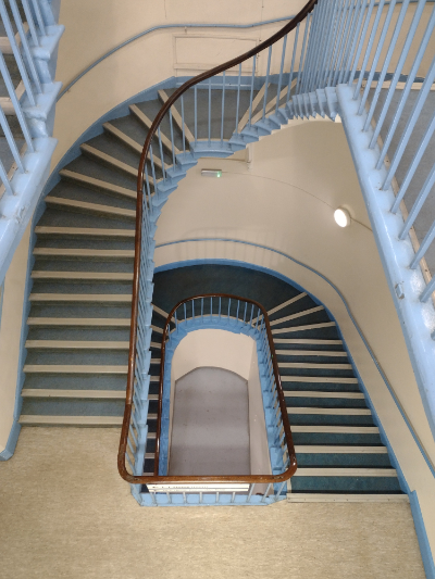 photograph of a set of internal stairs with a wooden banister and blue painted uprights, curving round two floors, steps are blue and landings beige