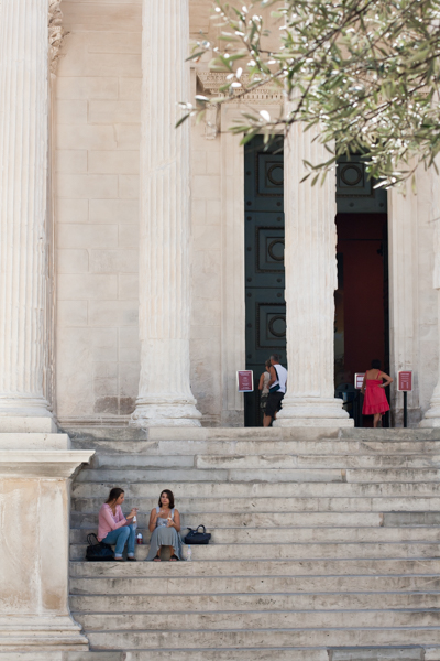 colour photograph, portrait layout, looking at a large set of marble steps with wide columns, two young ladies sitting on the left and some people entering the temple at the top of the steps