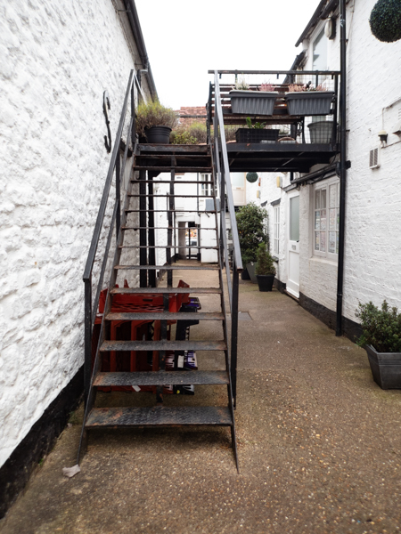 colour photograph, portrait layout, looking at a set of metal fire escape steps on the left with white building walls on either side and a metal footway leading from the top of the steps across to the right into the building on the right.  Another set of steps in the background and some plants on the footway at the top.