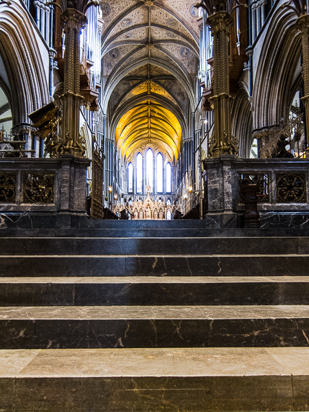 a set of stone steps leading up towards some marble altar rails. Beyond the rails, there are some stone arches on either side and a high ceiling.  At the far end in the centre of the picture is a brightly lit window.